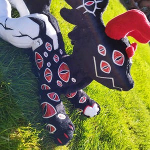 May include: A plush creature with a black body, red eyes, and red horns. The plush has white accents on its tail and legs. The creature is lying on green grass.