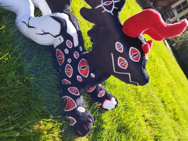 May include: A plush creature with a black body, red eyes, and red horns. The plush has white accents on its tail and legs. The creature is lying on green grass.