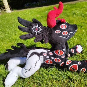 May include: A plush creature with black fur, red horns, and multiple red and white eye designs. The creature has white and black accents on its limbs and body. It is lying on a bed of green grass.