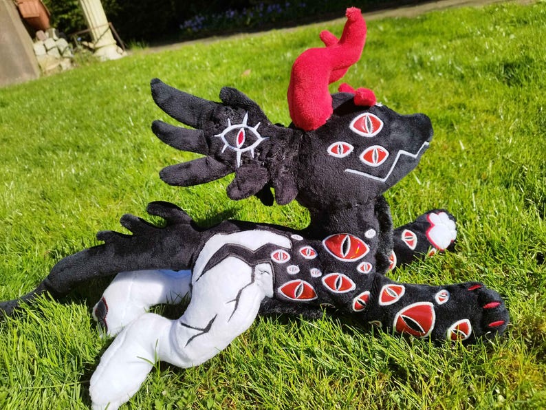 May include: A plush creature with black fur, red horns, and multiple red and white eye designs. The creature has white and black accents on its limbs and body. It is lying on a bed of green grass.