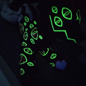 May include: Black plush creature with glowing green eye designs and accents. The eyes have red centers and are outlined in green. The creature's design glows in the dark, creating a striking visual effect.