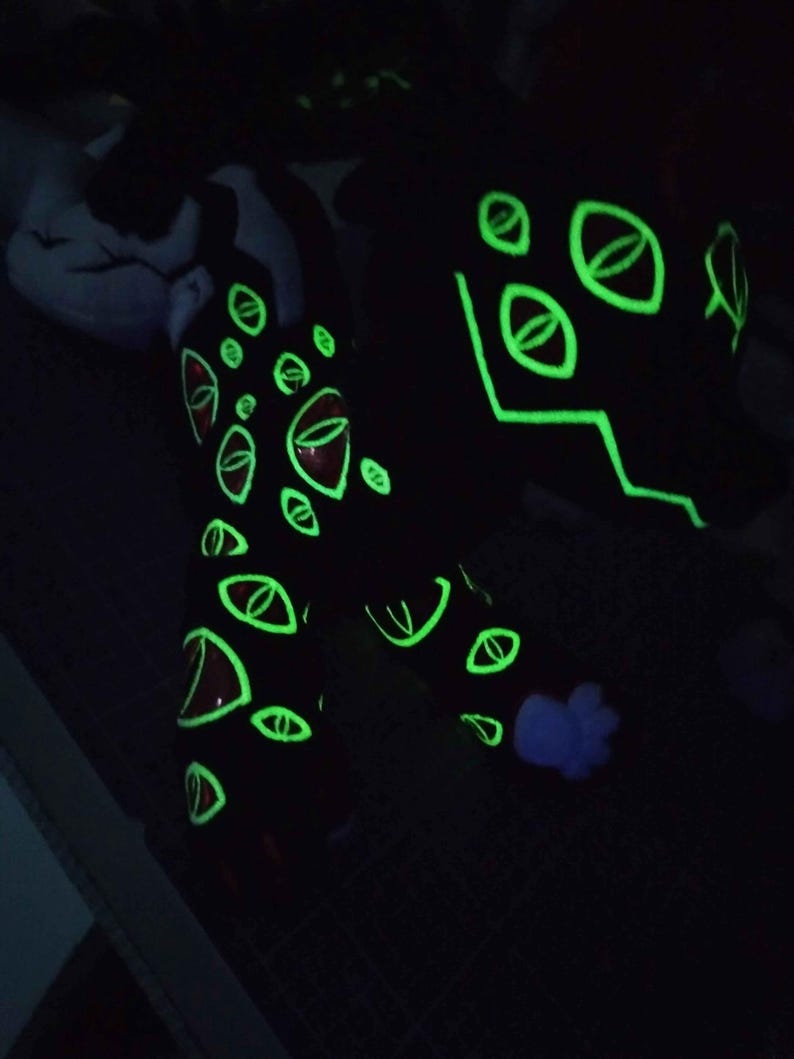 May include: Black plush creature with glowing green eye designs and accents. The eyes have red centers and are outlined in green. The creature's design glows in the dark, creating a striking visual effect.