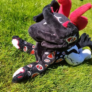 May include: A plush toy creature with a black body, red horns, and multiple red-and-white eyes. The toy is lying on green grass, with its legs and arms outstretched. The toy's design features a mix of black, red, white, and orange colors.
