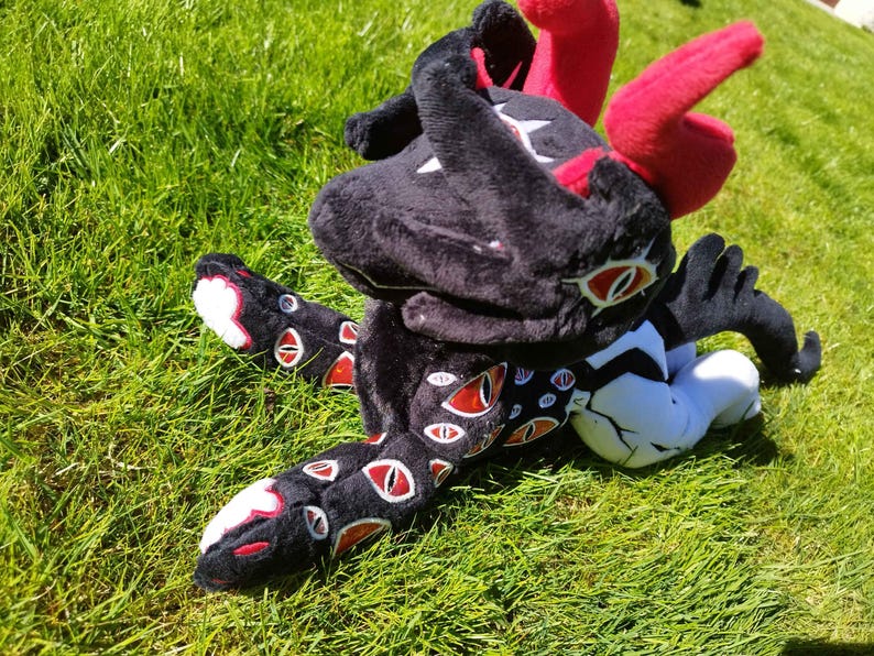 May include: A plush toy creature with a black body, red horns, and multiple red-and-white eyes. The toy is lying on green grass, with its legs and arms outstretched. The toy's design features a mix of black, red, white, and orange colors.