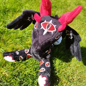 May include: A plush dragon toy with a black body, red horns, and wings. The dragon has white and red eye-like designs on its head and body. The toy is lying on green grass.