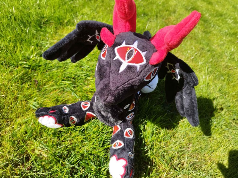 May include: A plush dragon toy with a black body, red horns, and wings. The dragon has white and red eye-like designs on its head and body. The toy is lying on green grass.