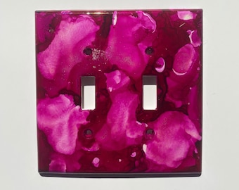 May include: A pink and white abstract design light switch cover with two toggle switches.