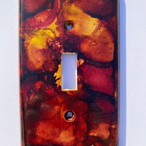 May include: A light switch cover with a red, orange, and yellow abstract design. The cover has a white switch plate.