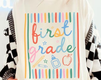 May include: An ivory t-shirt featuring a colourful graphic with the text "first grade" in cursive, surrounded by vertical stripes, a pencil, glue bottle, and apple icon. The shirt is paired with a red trucker hat with "MAD LOVE FOR YOU" text.