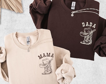 Puede incluir: Se muestran dos sudaderas de manga larga, una beige y otra marrón. La sudadera beige tiene la palabra "MAMA" y un gráfico de sombrero y bota de vaquero. La sudadera marrón tiene "DADA" y un gráfico similar. Ambas tienen un collar pequeño.