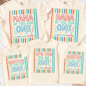 May include: Cream-colored t-shirts with colorful text and graphics. The shirts read "MAMA," "DADA," "BROTHER," "IT'S FUN TO BE," and "SISTER" with "ONE" and "Oliver's First Birthday." The shirts have vertical stripes.