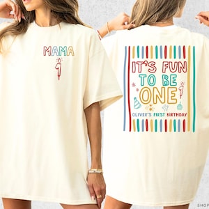 May include: Ivory-colored t-shirt with the word "MAMA" and a number "1" on the front. The back of the shirt features the text "IT'S FUN TO BE ONE" with birthday-themed graphics and "OLIVER'S FIRST BIRTHDAY".