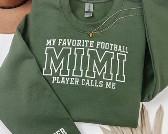 Könnte beinhalten: Ein olivgrünes Sweatshirt mit dem Text "MY FAVORITE FOOTBALL MIMI PLAYER CALLS ME" in weißen Blockbuchstaben. Der Ärmel hat den Namen "OLIVER" und ein Herz. Eine goldene Halskette und ein brauner Hut sind ebenfalls sichtbar.