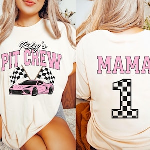 May include: Two white t-shirts with pink text and black and white checkered designs. One shirt says "Riley's Pit Crew" with a pink race car and checkered flags. The other shirt says "MAMA" with a large black and white checkered number "1".