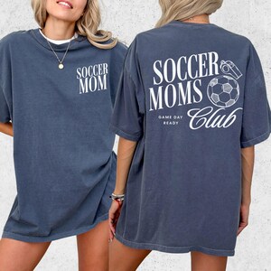 May include: A denim blue t-shirt with the words "SOCCER MOM" printed on the front. The back of the shirt features the text "SOCCER MOMS Club" with a soccer ball and whistle graphic. The words "GAME DAY READY" are also printed on the back.