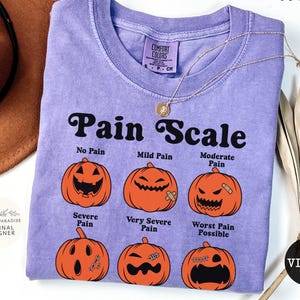 May include: A violet short-sleeved t-shirt featuring a black graphic titled Pain Scale. The design displays six orange pumpkins with varying facial expressions and labels: No Pain, Mild Pain, Moderate Pain, Severe Pain, Very Severe Pain, and Worst Pain Possible.