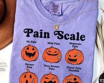 May include: A violet short-sleeved t-shirt featuring a black graphic titled Pain Scale. The design displays six orange pumpkins with varying facial expressions and labels: No Pain, Mild Pain, Moderate Pain, Severe Pain, Very Severe Pain, and Worst Pain Possible.