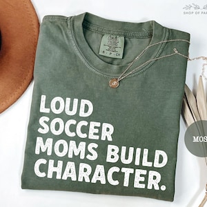 May include: An olive green t-shirt with the text "LOUD SOCCER MOMS BUILD CHARACTER" in white block letters. A gold necklace with a pendant rests on the shirt. The shirt is folded and displayed with a brown hat and dried plant.