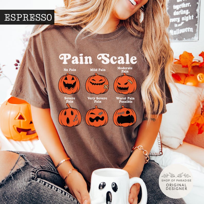 May include: An espresso-colored short-sleeve t-shirt featuring a graphic of six orange pumpkins with varying facial expressions labeled as a Pain Scale. The text reads No Pain, Mild Pain, Moderate Pain, Severe Pain, Very Severe Pain, and Worst Pain Possible.