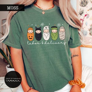 K&ouml;nnte beinhalten: Ein moosgr&uuml;nes Kurzarm-T-Shirt mit einer Grafik von f&uuml;nf in Windeln gewickelten Babys, die als K&uuml;rbis, Fledermaus, Geist, Frankensteins Monster und Mumie verkleidet sind. Der Text "labor & delivery" steht in wei&szlig;er Schreibschrift darunter.