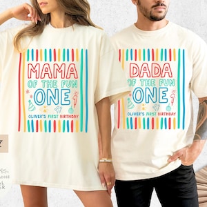 May include: Ivory-colored t-shirts with colorful text and graphics. One shirt reads "MAMA OF THE FUN ONE" and the other reads "DADA OF THE FUN ONE." Both shirts include "OLIVER'S FIRST BIRTHDAY" text and birthday-themed graphics.