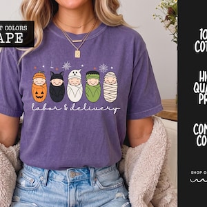 K&ouml;nnte beinhalten: Ein traubenfarbenes Kurzarm-T-Shirt mit einer Grafik von f&uuml;nf in Windeln gewickelten Babys, die als K&uuml;rbis, Fledermaus, Geist, Frankensteins Monster und Mumie verkleidet sind. Der Text "labor & delivery" steht in wei&szlig;er Schreibschrift darunter.