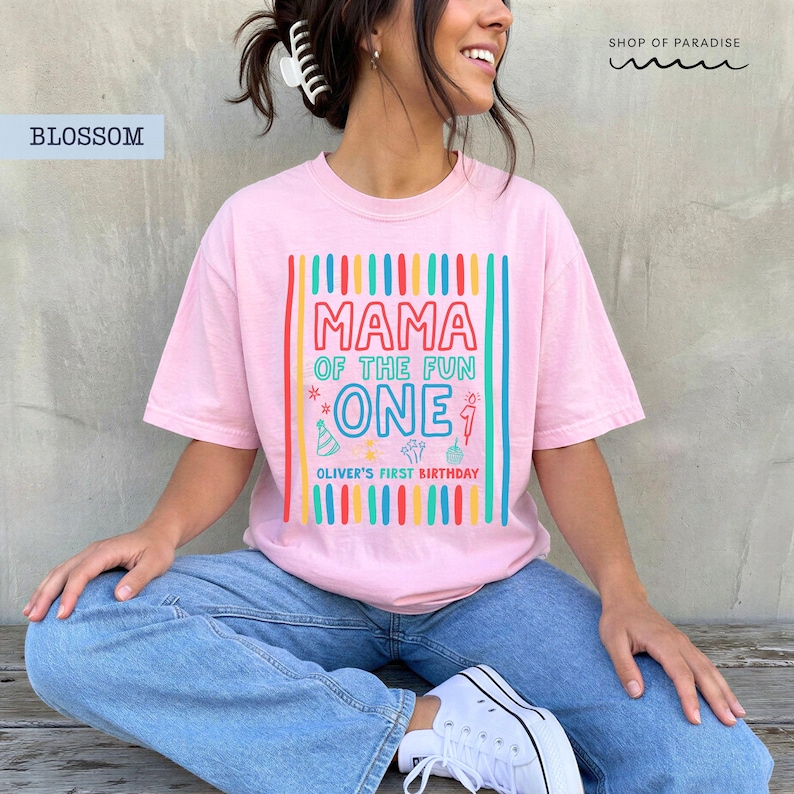 May include: A light pink t-shirt with the text "MAMA OF THE FUN ONE" in colorful letters. Below the text is "OLIVER'S FIRST BIRTHDAY". The shirt has a birthday theme with colorful vertical stripes.