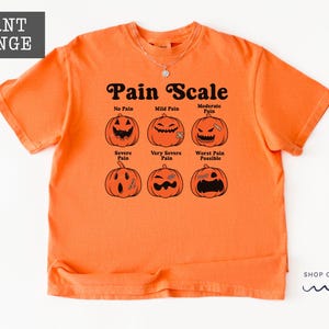 May include: A burnt orange short-sleeve t-shirt featuring a black graphic of six jack-o'-lanterns labeled as a Pain Scale. The text reads No Pain, Mild Pain, Moderate Pain, Severe Pain, Very Severe Pain, and Worst Pain Possible in black font.