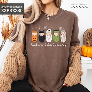 K&ouml;nnte beinhalten: Ein espressobraunes T-Shirt mit einer Grafik von f&uuml;nf in Windeln gewickelten Babys, die als K&uuml;rbis, Fledermaus, Geist, Frankenstein und Mumie verkleidet sind. Der Text "labor & delivery" steht unter den Babys, erg&auml;nzt durch Spinnennetze.