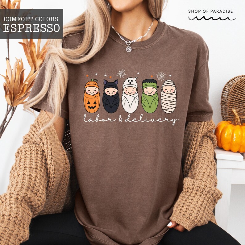 K&ouml;nnte beinhalten: Ein espressobraunes T-Shirt mit einer Grafik von f&uuml;nf in Windeln gewickelten Babys, die als K&uuml;rbis, Fledermaus, Geist, Frankenstein und Mumie verkleidet sind. Der Text "labor & delivery" steht unter den Babys, erg&auml;nzt durch Spinnennetze.