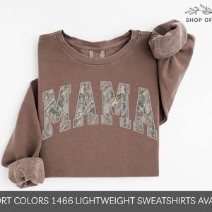 Puede incluir: Una sudadera marr&oacute;n con la palabra "MAMA" en un patr&oacute;n de camuflaje en el pecho. Las mangas est&aacute;n enrolladas y el texto "SHOP OF PARADISE" est&aacute; en la parte superior derecha. La parte inferior de la imagen dice "COMFORT COLORS 1466 LIGHTWEIGHT SWEATSHIRTS AVAILABLE!"
