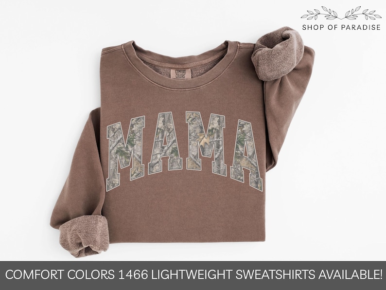 Puede incluir: Una sudadera marr&oacute;n con la palabra "MAMA" en un patr&oacute;n de camuflaje en el pecho. Las mangas est&aacute;n enrolladas y el texto "SHOP OF PARADISE" est&aacute; en la parte superior derecha. La parte inferior de la imagen dice "COMFORT COLORS 1466 LIGHTWEIGHT SWEATSHIRTS AVAILABLE!"