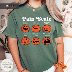 May include: A moss green short-sleeve t-shirt featuring a white graphic titled Pain Scale. The design displays six orange jack-o'-lanterns with varying facial expressions labeled No Pain, Mild Pain, Moderate Pain, Severe Pain, Very Severe Pain, and Worst Pain Possible.