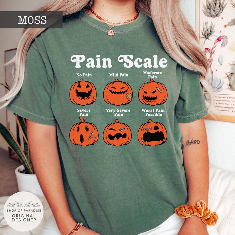 May include: A moss green short-sleeve t-shirt featuring a white graphic titled Pain Scale. The design displays six orange jack-o'-lanterns with varying facial expressions labeled No Pain, Mild Pain, Moderate Pain, Severe Pain, Very Severe Pain, and Worst Pain Possible.