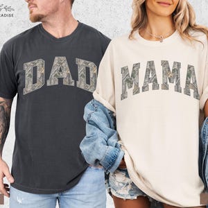 Puede incluir: Dos camisetas de manga corta, una color pimienta y otra color marfil. La palabra "DAD" est&aacute; impresa en la camiseta color pimienta, y la palabra "MAMA" est&aacute; impresa en la camiseta color marfil. Ambas palabras tienen un patr&oacute;n de camuflaje.