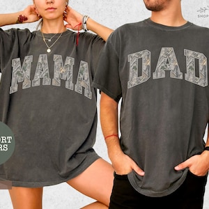 Puede incluir: Dos camisetas gris carb&oacute;n, una con "MAMA" y otra con "DAD" en un estampado de camuflaje. Las camisetas son de la colecci&oacute;n "Comfort Colors" y son de color "Pepper".