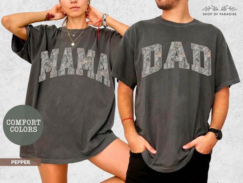 Puede incluir: Dos camisetas gris carb&oacute;n, una con "MAMA" y otra con "DAD" en un estampado de camuflaje. Las camisetas son de la colecci&oacute;n "Comfort Colors" y son de color "Pepper".