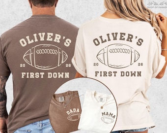 May include: Two t-shirts with a football design. The back of the shirts feature the text "OLIVER'S FIRST DOWN" with a football graphic and the numbers "20" and "26". One shirt is brown, the other is cream. Smaller shirts say "DADA" and "MAMA".