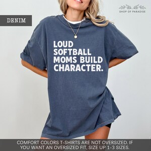 May include: Denim-colored t-shirt with the text "LOUD SOFTBALL MOMS BUILD CHARACTER" in white. The shirt is a relaxed fit and has short sleeves. The text "DENIM" is in a box in the upper left corner. The brand name "SHOP OF PARADISE" is in the upper right corner.