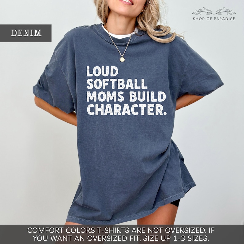 May include: Denim-colored t-shirt with the text "LOUD SOFTBALL MOMS BUILD CHARACTER" in white. The shirt is a relaxed fit and has short sleeves. The text "DENIM" is in a box in the upper left corner. The brand name "SHOP OF PARADISE" is in the upper right corner.