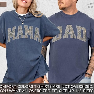 Puede incluir: Dos camisetas de color denim. Una dice "MAMA" y la otra "DAD" en una fuente de estilo camuflaje. Las camisetas forman parte de la colecci&oacute;n Comfort Colors. El logotipo de Shop of Paradise est&aacute; en la esquina superior derecha.
