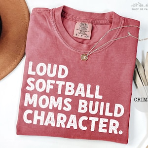 May include: Crimson-colored t-shirt with the text "LOUD SOFTBALL MOMS BUILD CHARACTER" in white block letters. A gold necklace with a pendant rests on the shirt. The shirt is a Comfort Colors brand.
