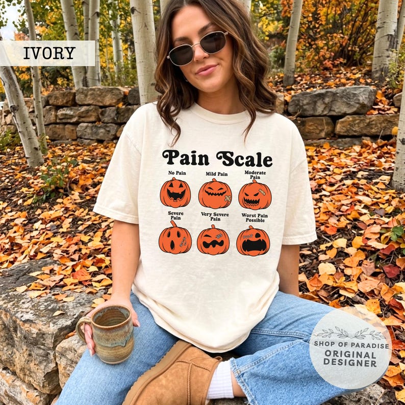 May include: An ivory t-shirt featuring a graphic of six orange jack-o'-lanterns arranged in two rows. The text reads Pain Scale, with labels for each pumpkin: No Pain, Mild Pain, Moderate Pain, Severe Pain, Very Severe Pain, and Worst Pain Possible.