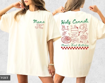 Può includere: T-shirt color avorio con testo e grafica verdi e rossi. Il davanti dice "Mama" con illustrazioni di pasta. Il retro dice "Holy Cannoli" con illustrazioni di cibo e "Oliver's First Birthday" con un bordo a scacchi.