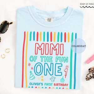May include: Light blue t-shirt with a colorful graphic that reads "MIMI OF THE FUN ONE" with additional text "OLIVER'S FIRST BIRTHDAY". The design includes vertical stripes and birthday-themed illustrations. Sunglasses and accessories are in the background.