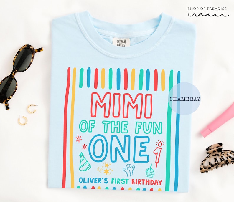 May include: Light blue t-shirt with a colorful graphic that reads "MIMI OF THE FUN ONE" with additional text "OLIVER'S FIRST BIRTHDAY". The design includes vertical stripes and birthday-themed illustrations. Sunglasses and accessories are in the background.