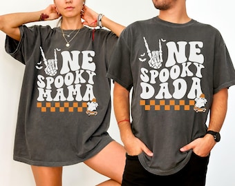 Puede incluir: Dos camisetas gris carbón con texto y gráficos en blanco. Una dice ONE SPOOKY MAMA y la otra ONE SPOOKY DADA. Ambas incluyen una mano de esqueleto haciendo un signo de rock, murciélagos pequeños y un fantasma sobre un monopatín con un patrón ajedrezado.