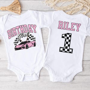 May include: Two white baby onesies with a pink race car theme. The front onesie says "Birthday Girl" with a pink race car and checkered flags. The back onesie says "Riley" with a checkered flag number one.