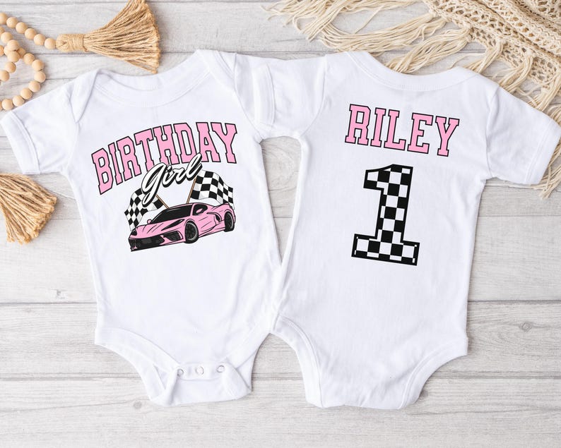 May include: Two white baby onesies with a pink race car theme. The front onesie says "Birthday Girl" with a pink race car and checkered flags. The back onesie says "Riley" with a checkered flag number one.
