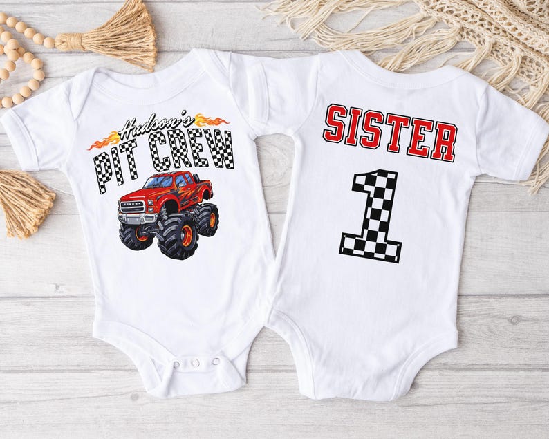 Pu&ograve; includere: Due body bianchi per neonato. Un body ha una grafica di un camion mostro rosso con il testo "Hudson's Pit Crew". L'altro body ha il testo "Sister" e il numero "1" con un motivo a scacchi.